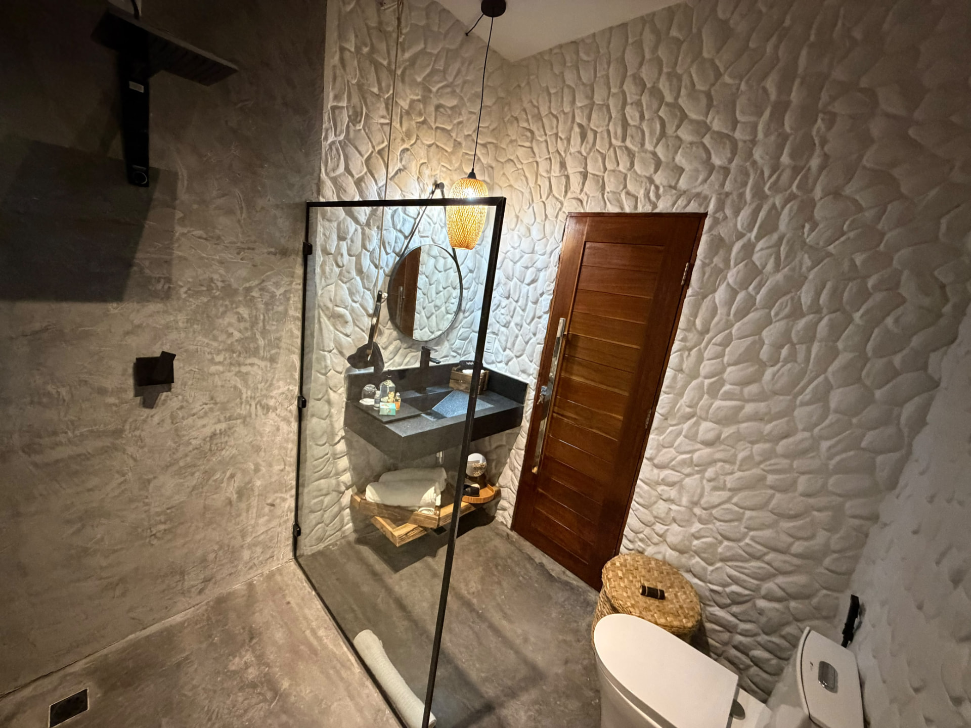Banheiro com box de vidro, luminária de palha e porta de madeira